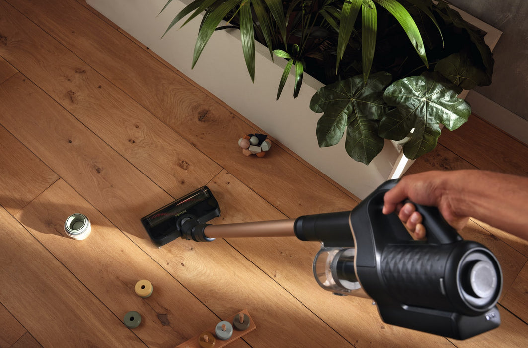 Person using a Miele Duoflex HX1 Cordless Stick Broom Vacuum Cleaner Total Care, Black & Gold SQLL0 on a wooden floor with toys scattered around. Capital Vacuum Raleigh Cary NC