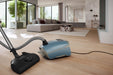 Miele Guard L1  Nordic Blue vacuum cleaner on a wooden floor with a modern living room in the background