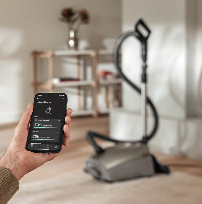 Person holding a smartphone with app controls for a Miele Titanium vacuum cleaner in a living room. Capital Vacuum Raleigh Cary NC