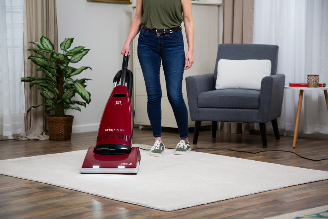 Woman using a Riccar R21 Spirit Plus red vacuum cleaner on a wooden floor with a chair and plant in the background. Riccar R21 Spirit Plus Upright Vacuum Capital Vacuum Raleigh Cary NC