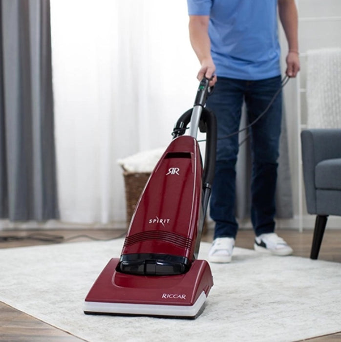Person using a red Riccar Spirit vacuum cleaner on a carpeted floor. Riccar R21 Spirit Upright Vacuum Capital Vacuum Raleigh Cary NC