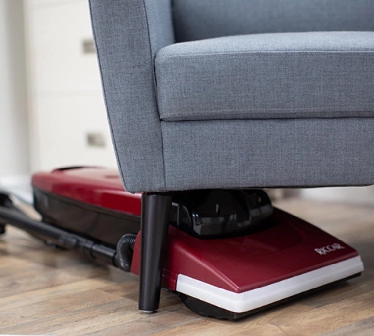 Riccar red and white vacuum cleaner under a gray sofa on a wooden floor. Riccar R21 Spirit Upright Vacuum Capital Vacuum Raleigh Cary NC