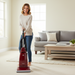 Woman using a Riccar R21 vacuum cleaner in a living room with a gray sofa and coffee table.