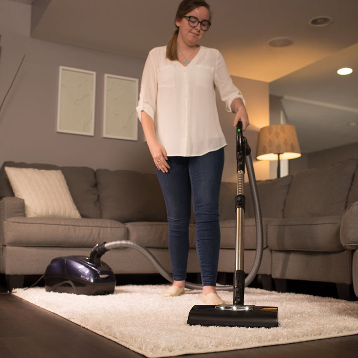 Woman using a Riccar Prima vacuum cleaner on a carpet in a living room. Riccar Prima canister vacuum cleaner with compact nozzle. Capital Vacuum Raleigh Cary NC