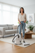 A woman stands in a bright living room holding a white and gray canister Sebo vacuum cleaner on a light rug in front of a gray sofa.